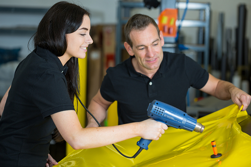 Male and female technicians applying yellow vinyl to vehicle . Female is holding dryer